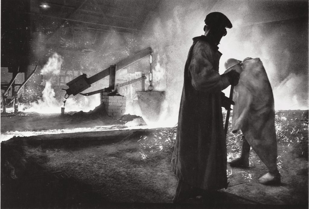 Black and white photograph of two men in long coats and hats in front of flames, sparks, and smoke in an industrial setting. 