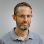 Headshot of white male with grey beard, short hair, and glasses in a grey polo shirt.