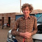 Photo of white male with curly strawberry blonde hair in a patterned brown button-down shirt, leaning against the hood of a truck. In the background is a brown building and two figures in the doorway.