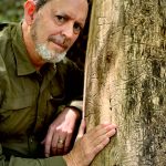 Picture of a white man with gray beard and short hair in a green long-sleeve shirt and standing next to tree, with his hand on the trunk.