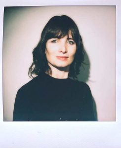 A polaroid photograph of the poet Lindsay Turner in front of a white background. She has shoulder-length dark brown hair and bangs and is wearing a dark sweater.