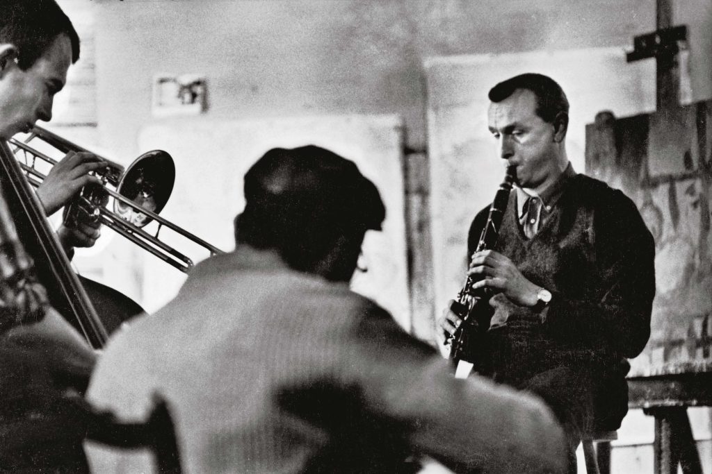 Black and white photograph of three men. Two men at the sides are playing wind instruments, and a seated man in the center is viewed from behind. 