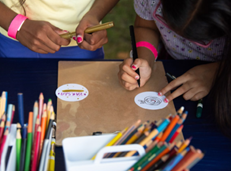 Photo 1: Two people decorate circular paper designs with colored pencils and markers at a table filled with art supplies.
Photo 2: A person uses a button-making press at a craft table covered with metal button parts, while others work in the background.
Photo 3: A colorful display of round buttons featuring animal illustrations and the text “South Side Science Festival” in orange, green, and blue designs.
