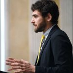 Person standing at a podium, captured in profile view, wearing a dark suit jacket, light patterned shirt, and a bright yellow tie. The individual’s hands are raised mid-gesture, suggesting they are speaking or presenting. The background shows tall columns and a neutral-toned wall, indicating an indoor formal setting.