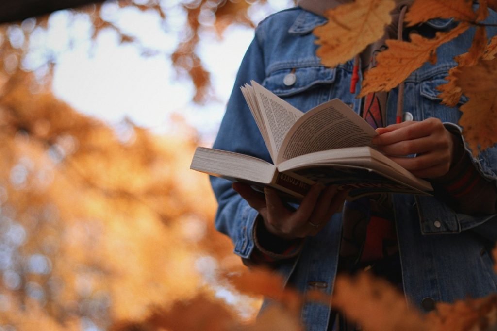 Hands holding an open book against a backdrop of vibrant orange autumn leaves. The reader wears a blue denim jacket, adding to the cozy, seasonal vibe.