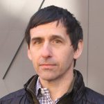 A photograph of Jonathan Thirkield, who has short dark hair and is wearing a blue and white shirt layered under a jacket. He is standing outdoors in front of a light brown background. 
