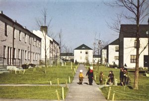 Children play and walk along a wide path lined with young trees and grassy lawns between rows of apartment buildings on a sunny day in a residential neighborhood. The scene gives a sense of community and outdoor activity.