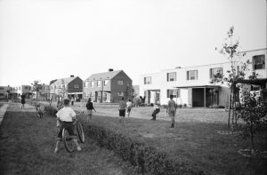 Black-and-white photo of a mid-20th-century suburban neighborhood. Several children are playing in an open grassy area between rows of modern-style houses. Some children are running, while others stand near bicycles. The houses in the background include brick and white facades, with large windows and minimal landscaping. A few young trees and shrubs line the open space, creating a sense of community and outdoor play.