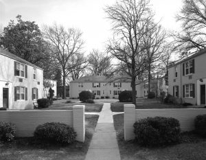 Historic black-and-white photograph showing a wide river with a steamboat traveling along it. The steamboat has a large paddle wheel at the rear and multiple decks with railings. In the background, there are tree-lined banks and a calm sky, giving the image a vintage Mississippi River setting.